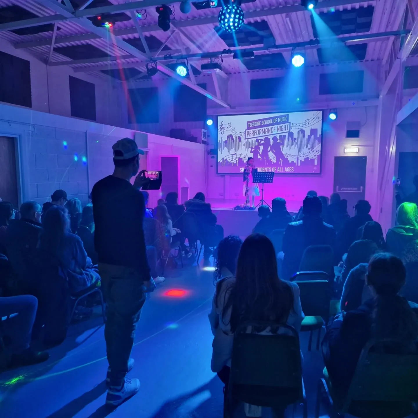 A young singer performing on the hall stage at a student performance night, with family and friends watching